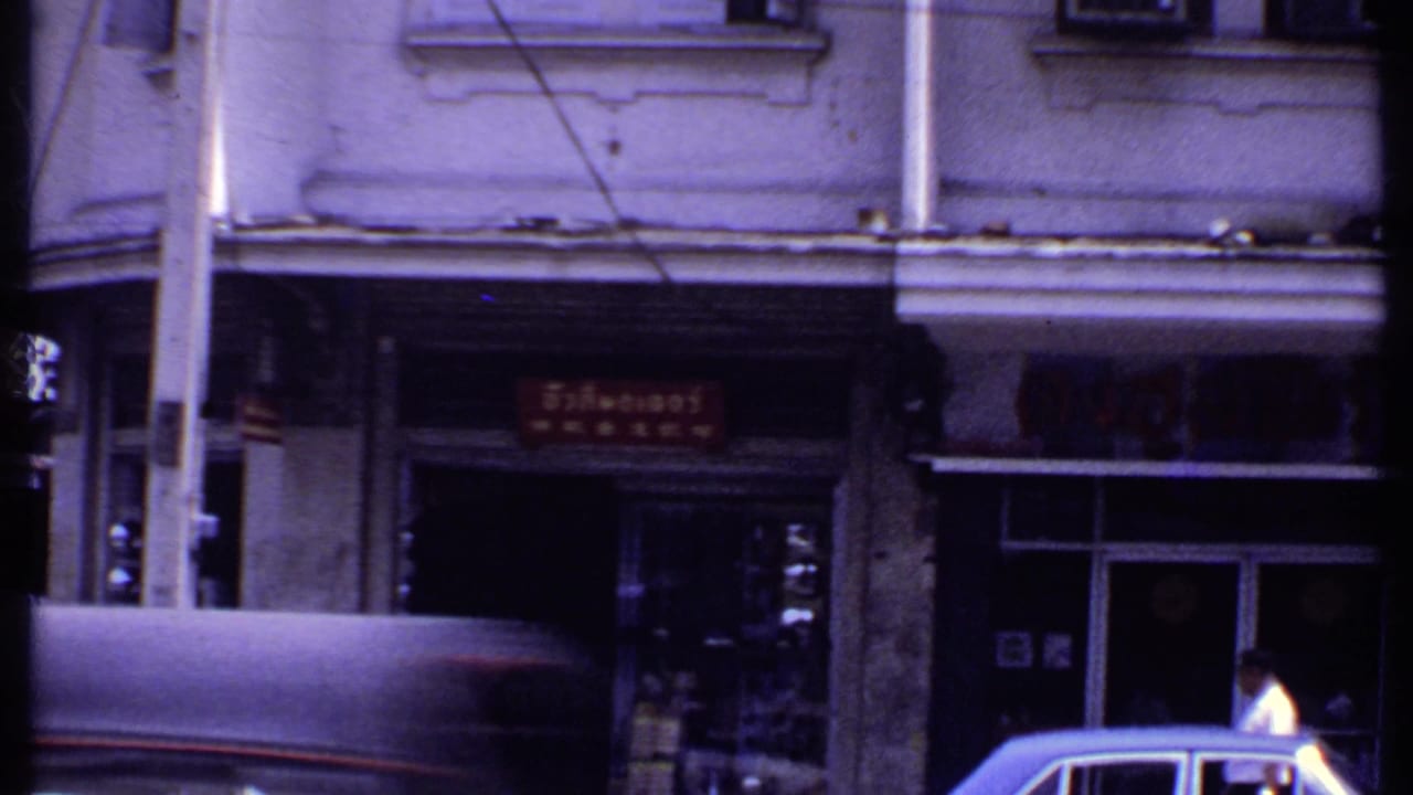 Archival street frame with a covered vehicle, pedestrians, red storefront signage, poles, and shaded shop entrances.