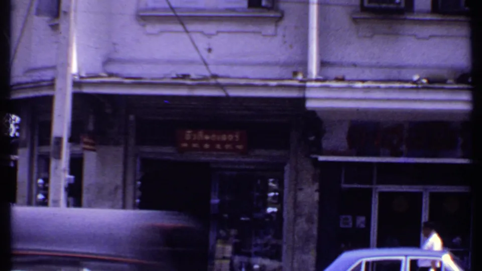 Archival street frame with a covered vehicle, pedestrians, red storefront signage, poles, and shaded shop entrances.