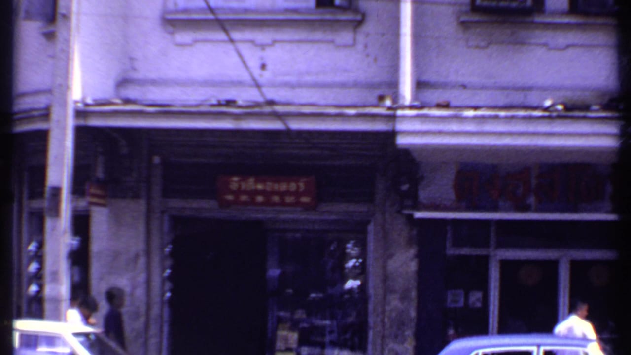 1969 street-level view of a red shop sign, shaded entrance, pedestrians, a parked car edge, and weathered building details.