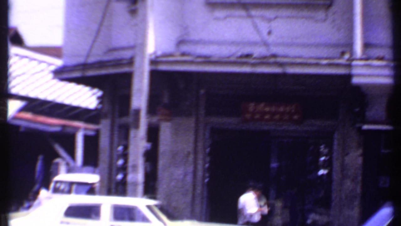 Archival urban frame showing a white classic car, pedestrians, shopfront signage, an awning, and a vertical street pole.