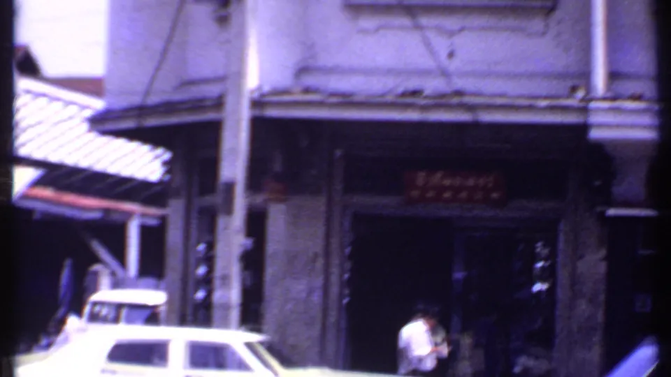 Archival urban frame showing a white classic car, pedestrians, shopfront signage, an awning, and a vertical street pole.