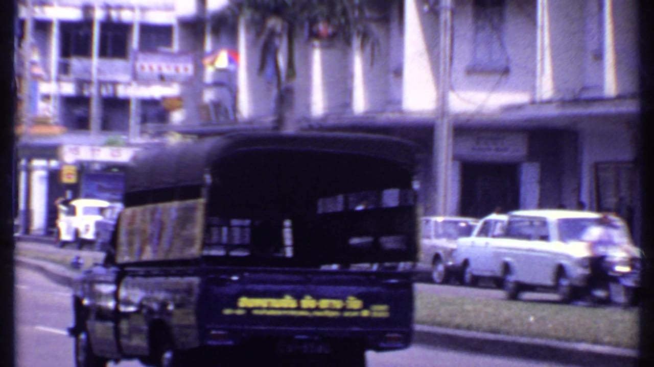 Auto rickshaw and older cars pass storefronts on a palm-lined street.