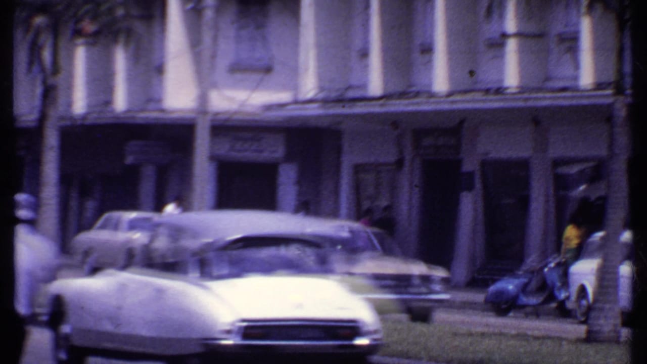 1969 India street scene showing an older white car, parked sedans, palm trunks, and arcaded commercial facades.