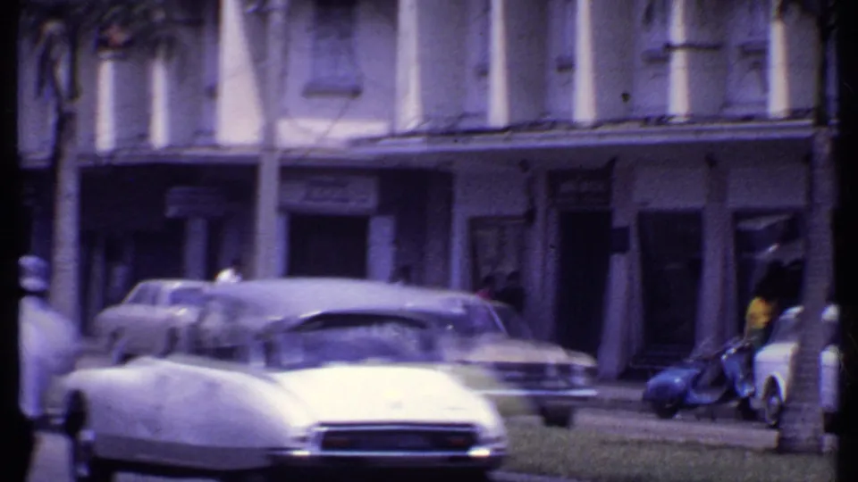 1969 India street scene showing an older white car, parked sedans, palm trunks, and arcaded commercial facades.