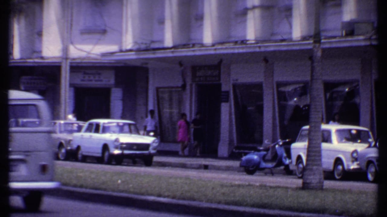 1969 India commercial street with older cars, scooters, pedestrians, palm trunks, and shopfronts seen in soft 8mm color.