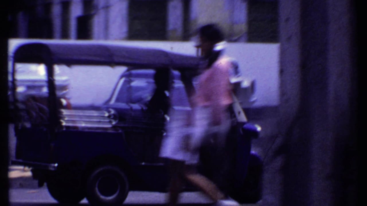 1969 India street view with a blurred pedestrian passing an auto rickshaw, parked cars, and a shaded building edge.