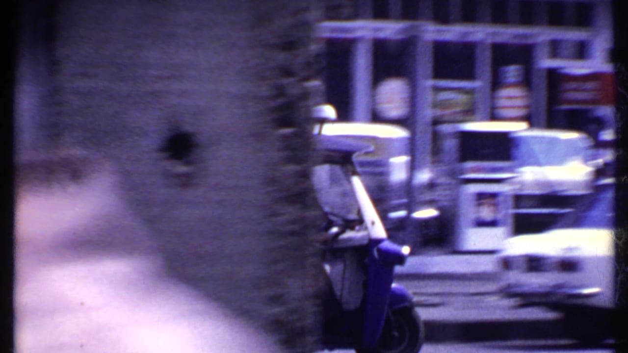 Blue three-wheeled vehicle and parked cars beside a storefront on a town street.