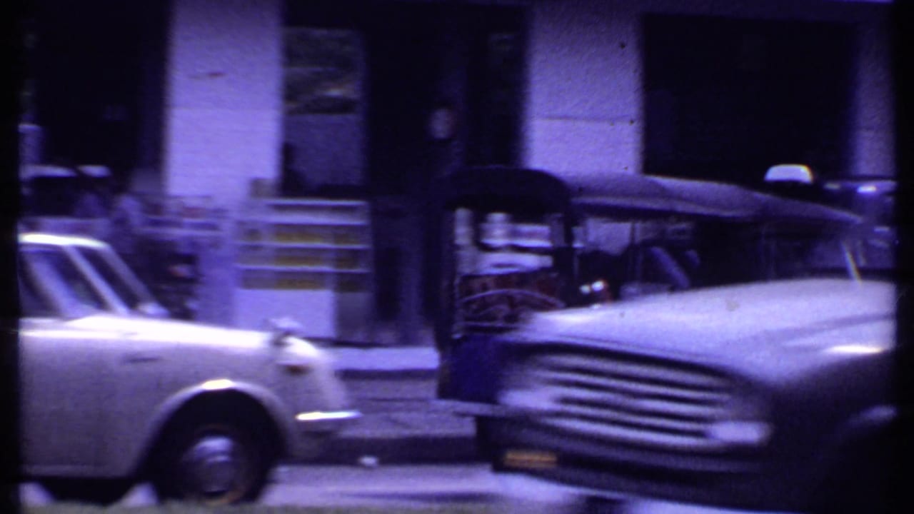 White sedan and larger road vehicle pass along a town street in 1969 India, with shopfronts and steps behind them.