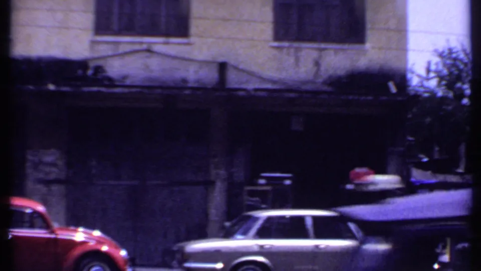 Town street frame from 1969 India showing a red car, a light-colored station wagon, and a darkened building frontage.