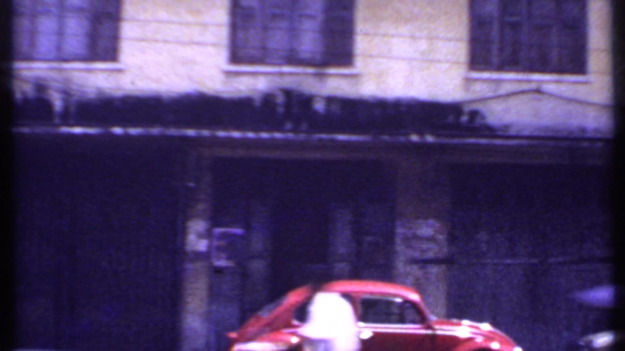 Red car on a 1969 India town road, with a dark doorway, barred upper windows, and soft purple 8mm color shift.