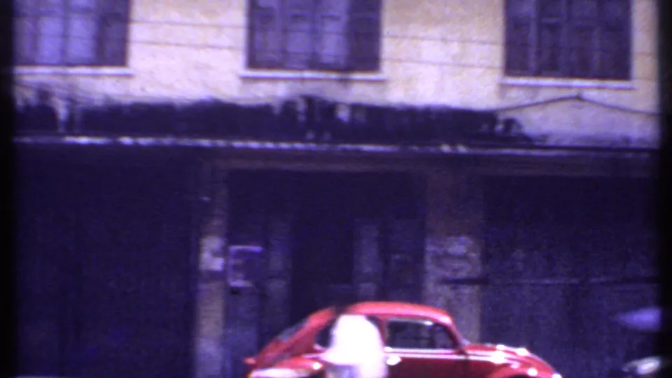 Red car on a 1969 India town road, with a dark doorway, barred upper windows, and soft purple 8mm color shift.