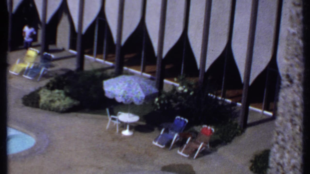 Elevated view of a hotel poolside with lounge chairs, umbrella, shrubs and tall facade columns.