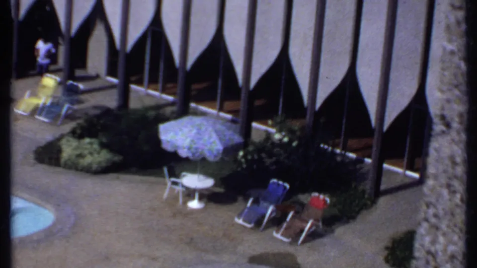 1969 India 8mm frame showing an elevated hotel poolside view with lounge chairs, a patterned umbrella, landscaping and vertical facade columns.