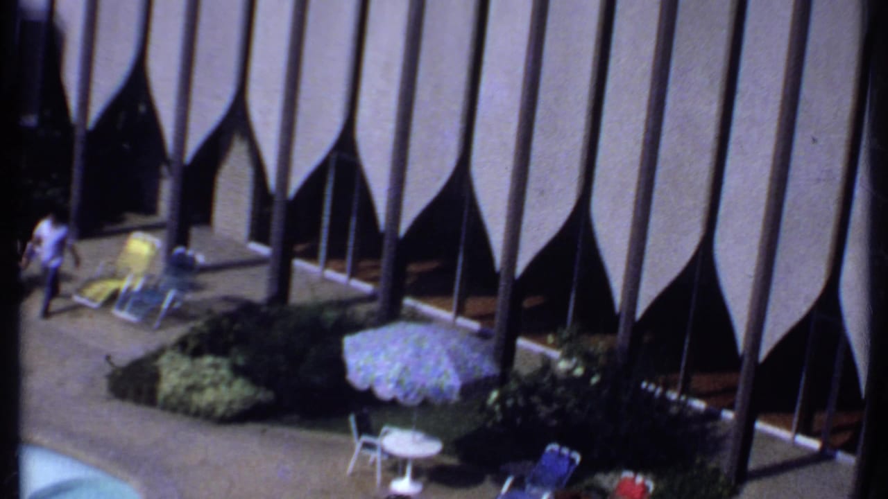 Person walking near lounge chairs and a patterned umbrella beside a hotel pool area.