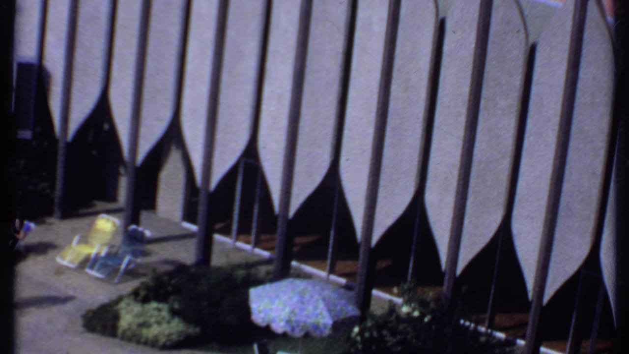 1969 India 8mm frame showing poolside chairs, a patterned umbrella, landscaping and partial figures near the swimming pool.