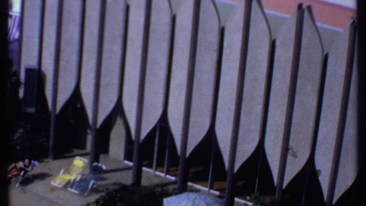 1969 India 8mm frame showing people resting near yellow lounge chairs, a patterned umbrella and tall scalloped facade panels.