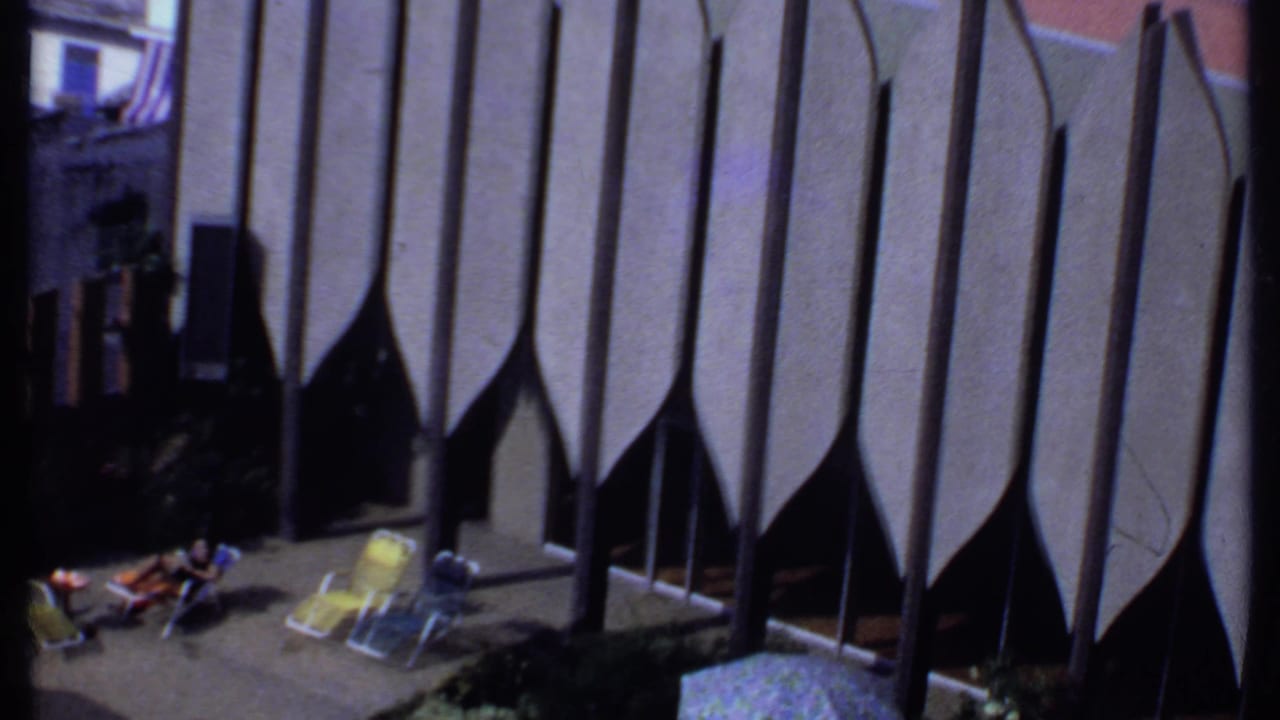 1969 India home-movie frame of a woman relaxing in a sunlit courtyard with folding chairs and strong architectural shadows.