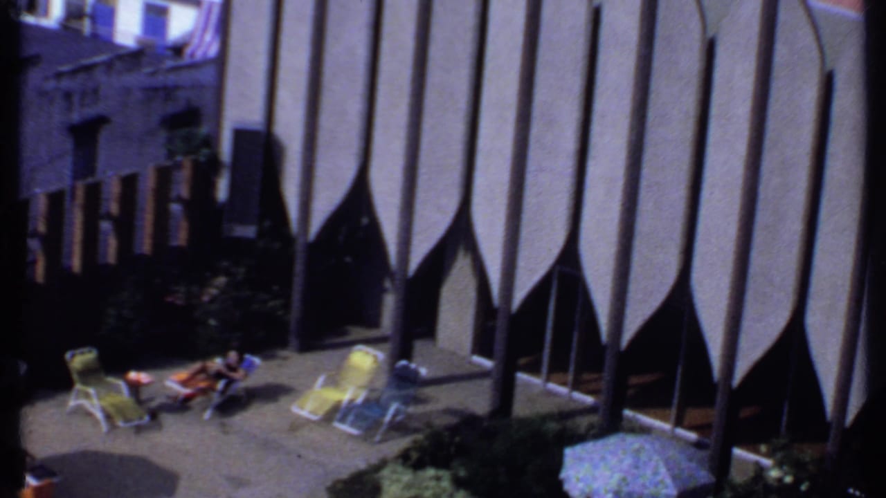 Courtyard view with a reclining woman, yellow chairs, shrubs, and tall vertical wall elements.