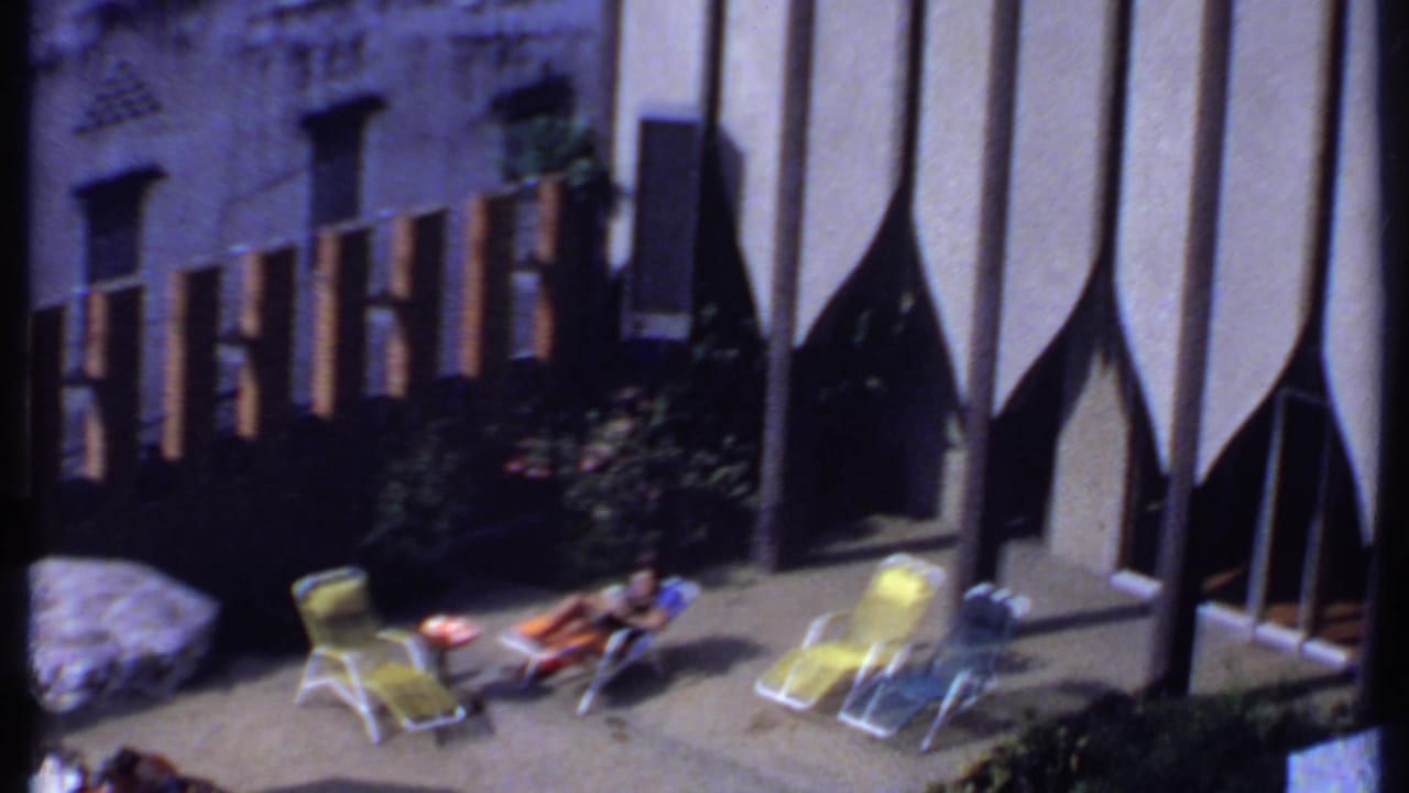 1969 India home-movie frame with a patterned patio umbrella, yellow chairs, and a woman reclining in the sunlit courtyard.