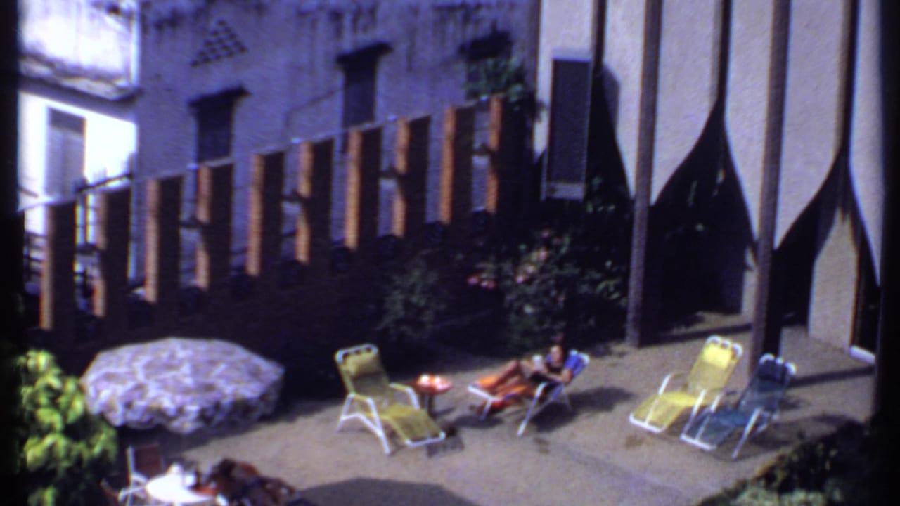 1969 India frame of a quiet courtyard leisure scene with a woman reclining, folding chairs, umbrella, shrubs, and vertical facade fins.