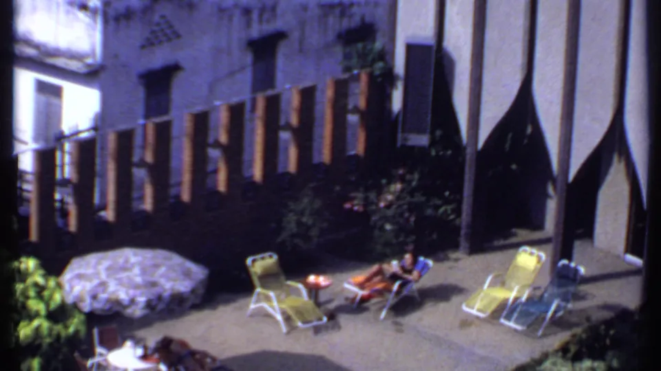 1969 India frame of a quiet courtyard leisure scene with a woman reclining, folding chairs, umbrella, shrubs, and vertical facade fins.