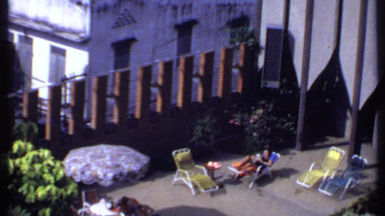 1969 India home-movie frame showing a woman reclining with reading material in a sunlit backyard beside a patterned umbrella and wooden fence.