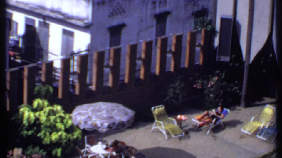 1969 India backyard scene with a woman reclining in a lounge chair near a small table, patterned umbrella, greenery, and wooden slat fence.