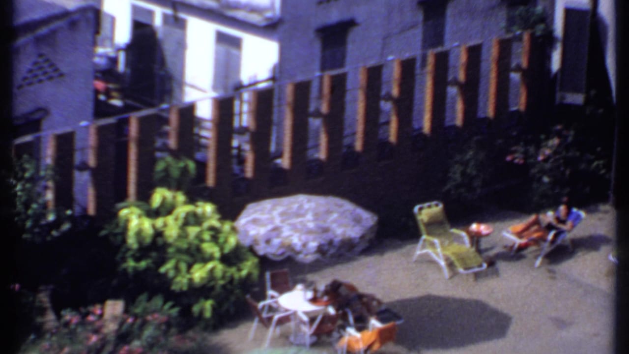 1969 India 8mm frame of a sunlit backyard patio, with lounge chairs, a patterned umbrella, plants, and a woman reclining near the fence.