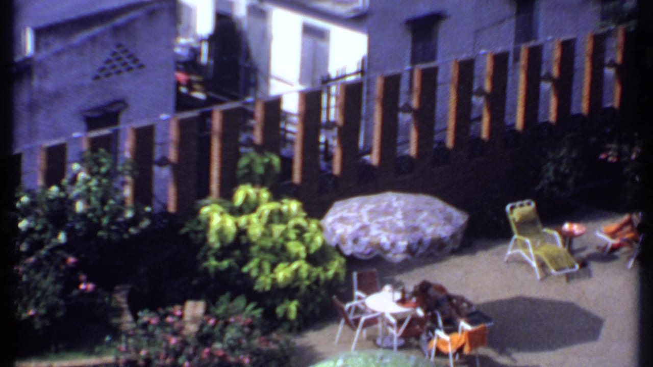 1969 India home-movie view of a woman reclining in a backyard patio area with a patterned umbrella, white table, plants, and slatted fence.