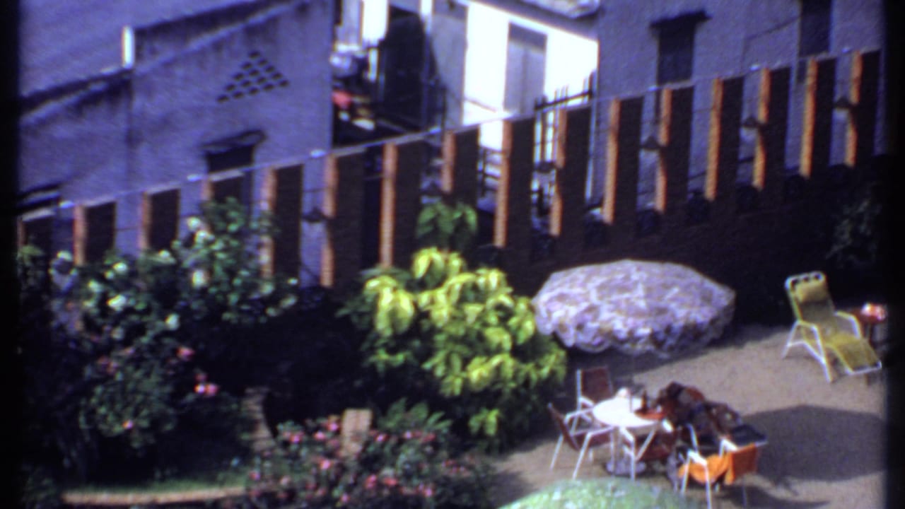 Sunlit patio with umbrellas, garden plants, chairs, and people seated near a pool area.