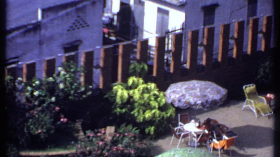 1969 India home-movie frame showing a sunlit patio with garden planting, umbrellas, chairs, and people seated near a pool area.