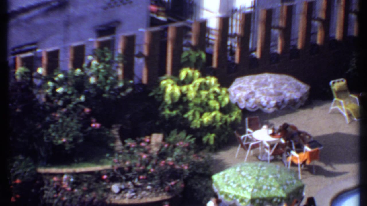 1969 India poolside patio scene with lush planting, patterned umbrellas, white tables, orange chairs, and seated people.