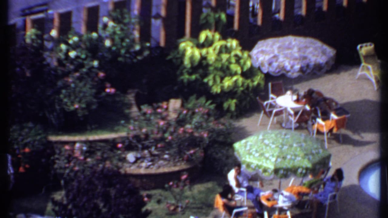 1969 India home-movie view of people seated around small tables on a planted outdoor patio with umbrellas and poolside chairs.