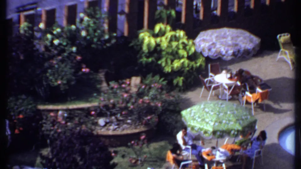 1969 India home-movie view of people seated around small tables on a planted outdoor patio with umbrellas and poolside chairs.