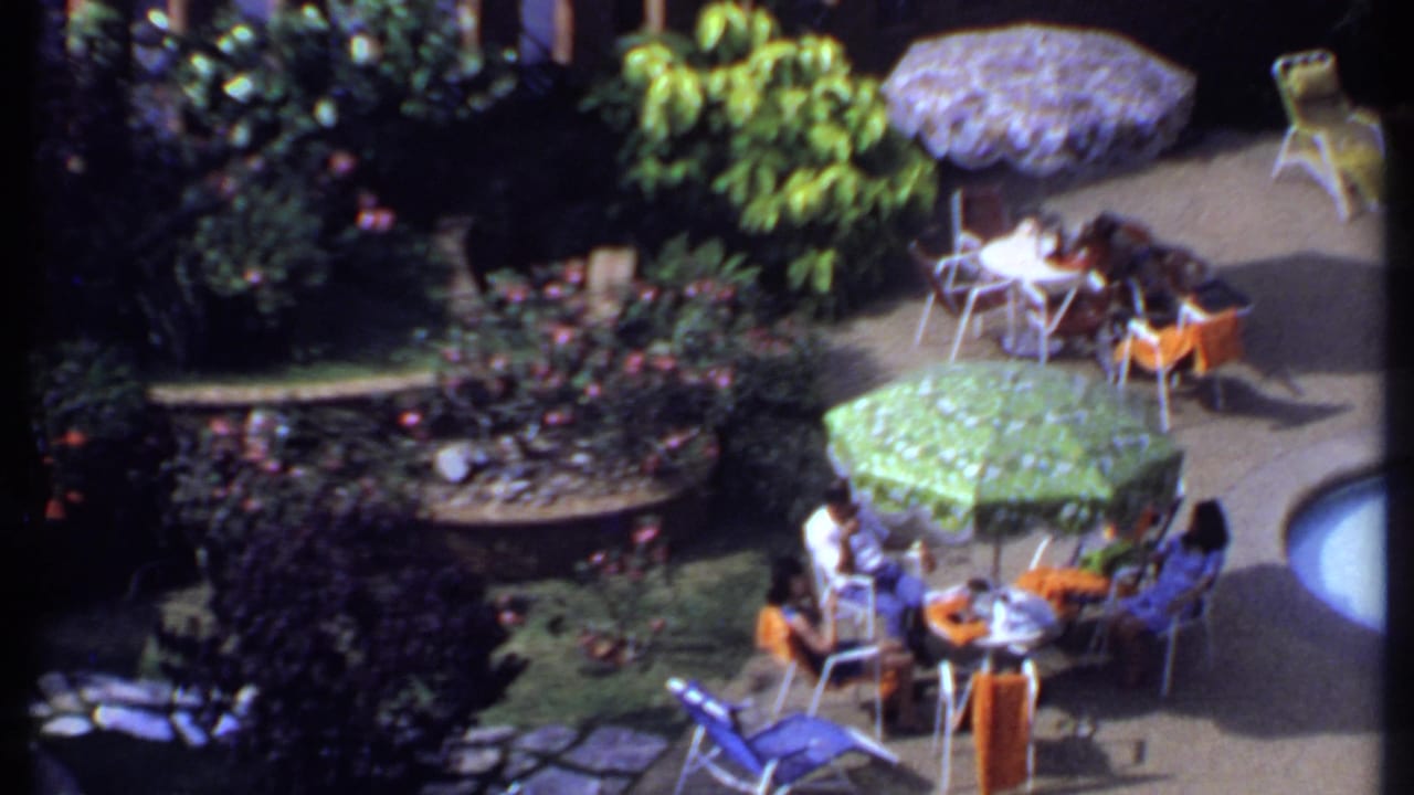 1969 India footage frame showing a poolside patio gathering with a green umbrella, orange chairs, white tables, and garden beds.
