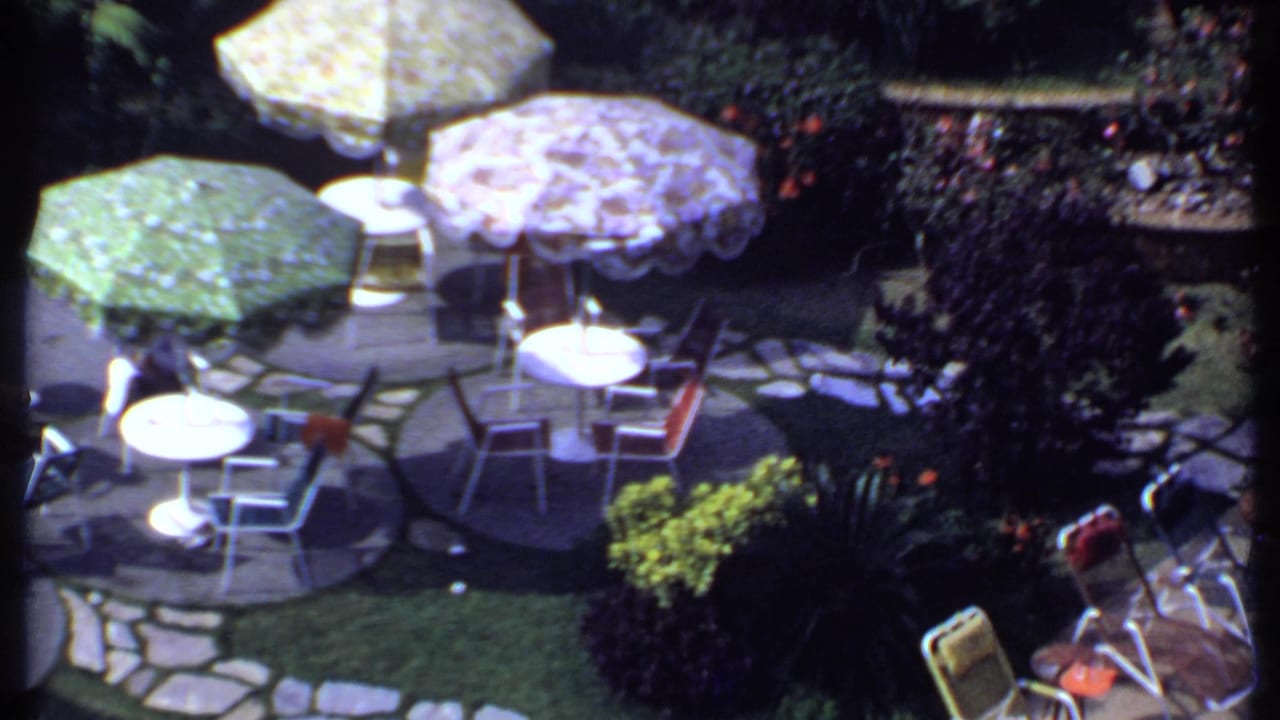 Patio with patterned umbrellas, white tables, metal chairs, stone paving, and surrounding greenery.