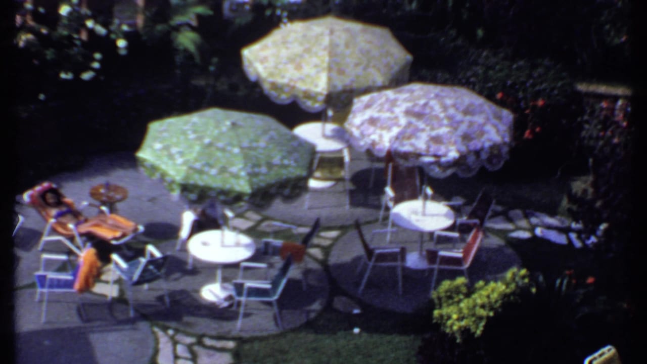 Person reclining on a patio chair near tables, chairs, and patterned umbrellas.