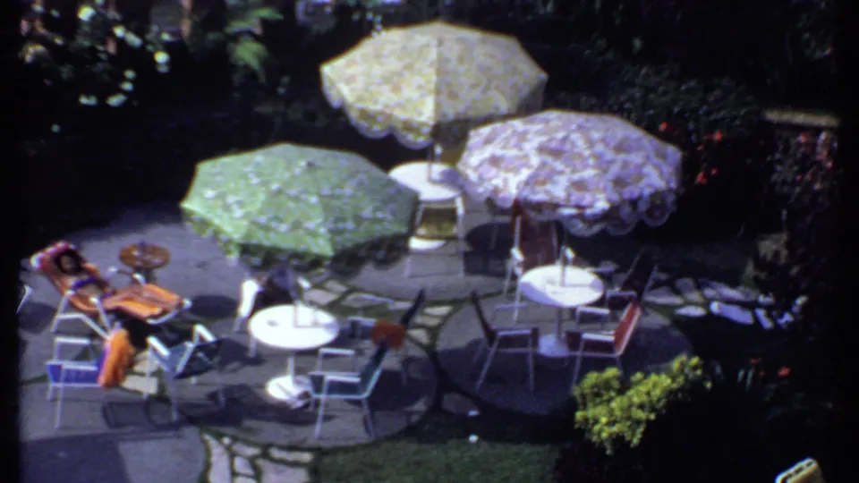 1969 India home-movie frame with a person relaxing beside patio tables and floral umbrellas.