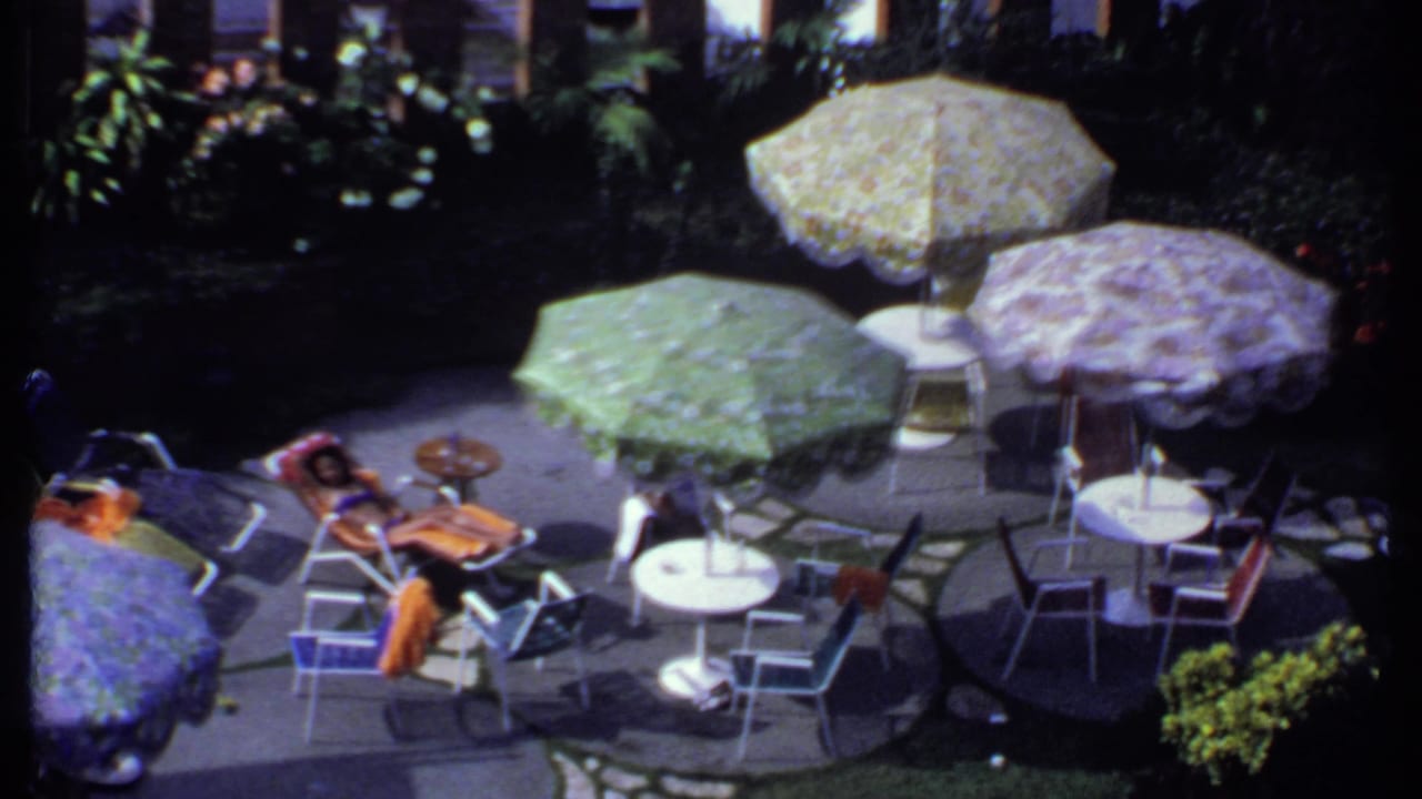 1969 India 8mm view across a shaded patio with floral umbrellas, seating, and a person resting nearby.