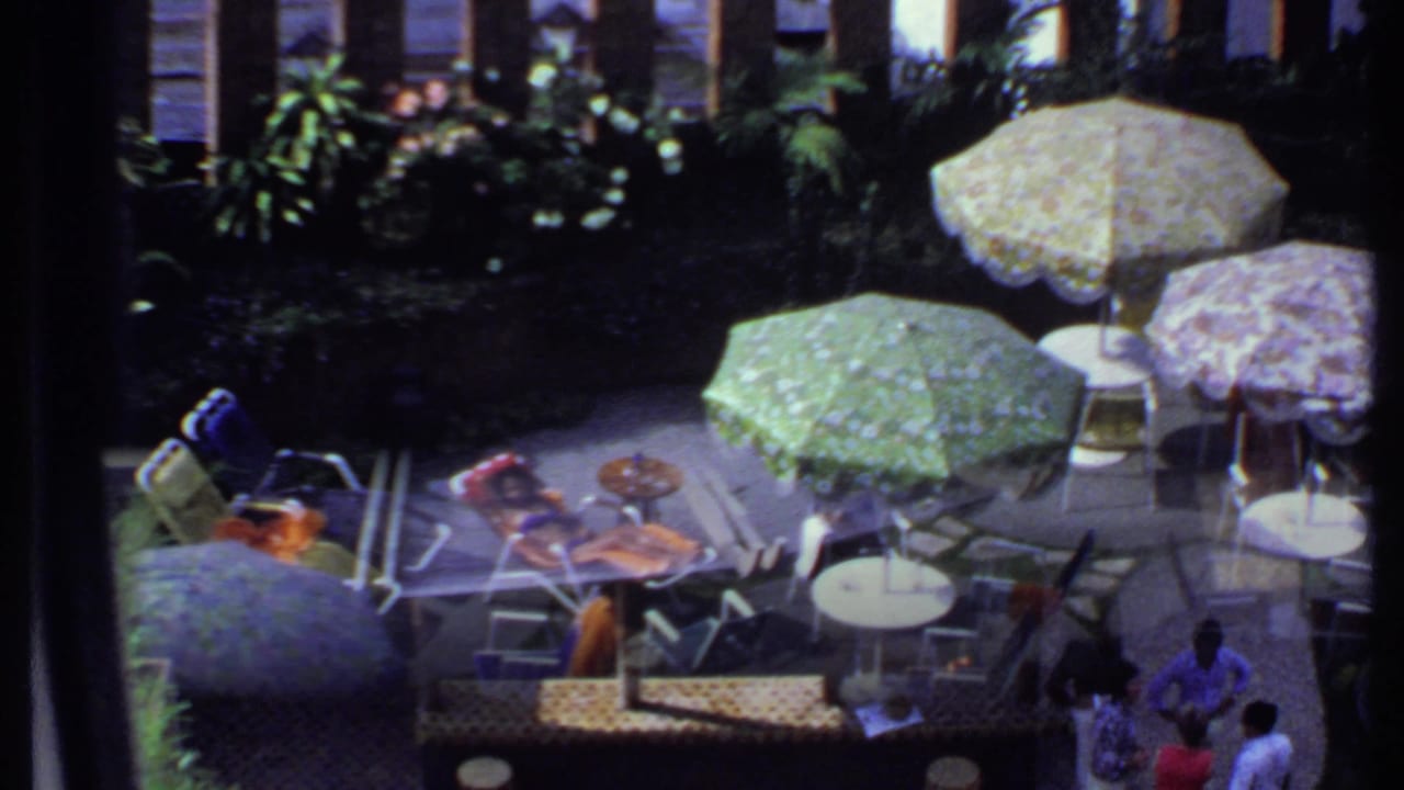 1969 India frame showing patterned patio umbrellas, chairs, garden plants, and people at the edge of the scene.
