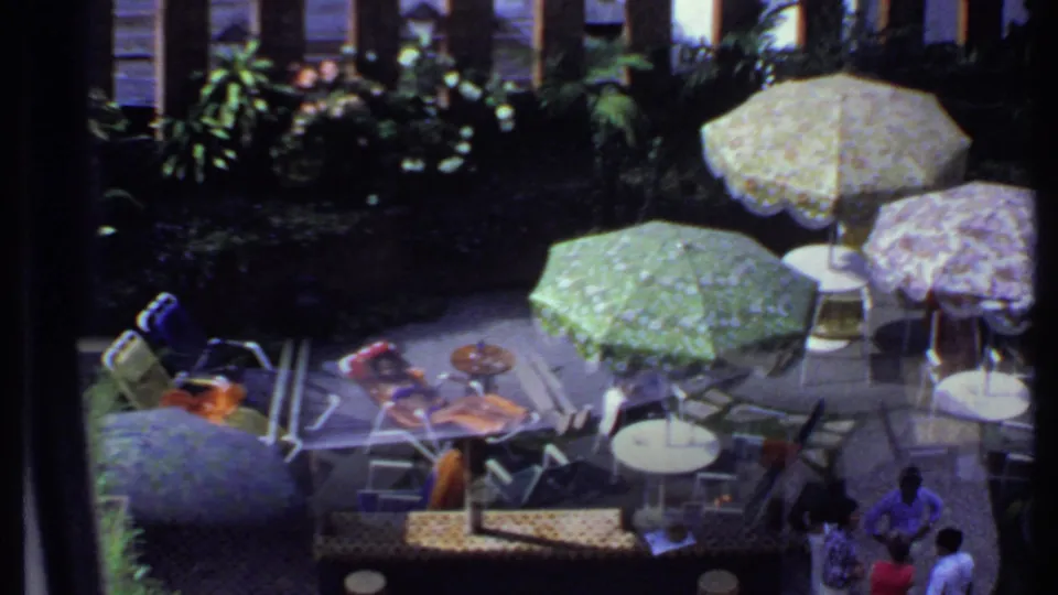 1969 India frame showing patterned patio umbrellas, chairs, garden plants, and people at the edge of the scene.
