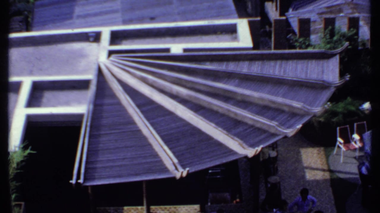 Elevated 1969 India home-movie view of the fan-shaped roof, nearby chairs, greenery, and small figures at the edge of the patio.