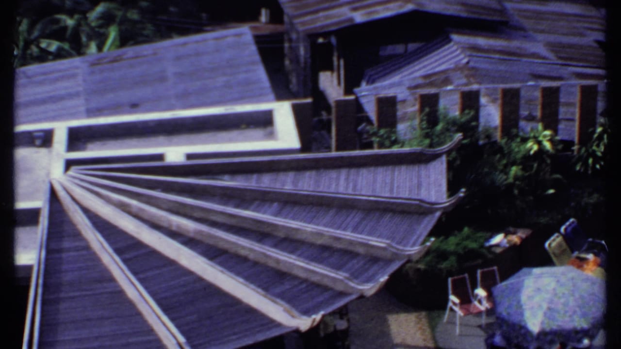 Final 1969 India frame with the fan-shaped roof, adjacent buildings, patio chairs, greenery, and small figures partly visible below.