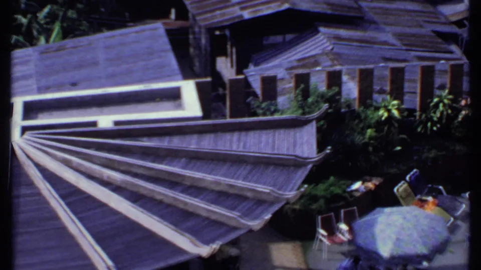 1969 India home-movie view over a sunlit terrace with tiered roofs, greenery, parasols, deck chairs, and tables.