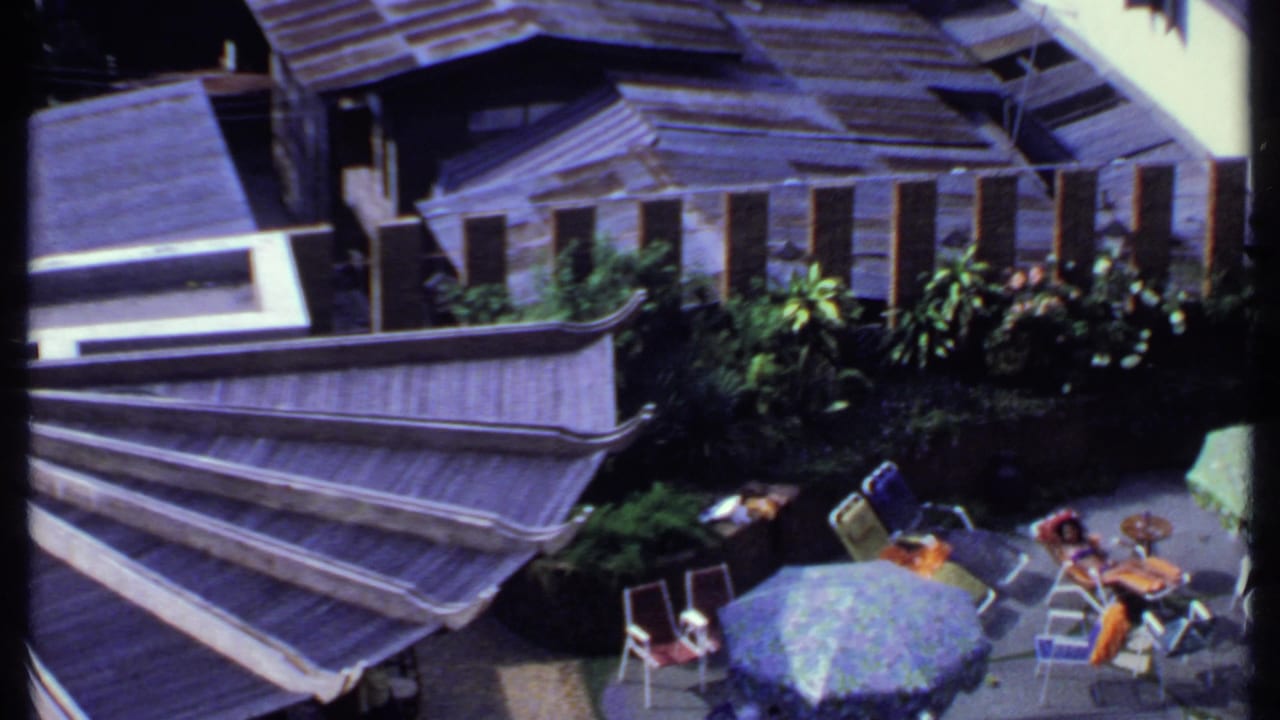 1969 India terrace scene with patterned parasols, metal deck chairs, small round tables, plants, and adults relaxing outdoors.