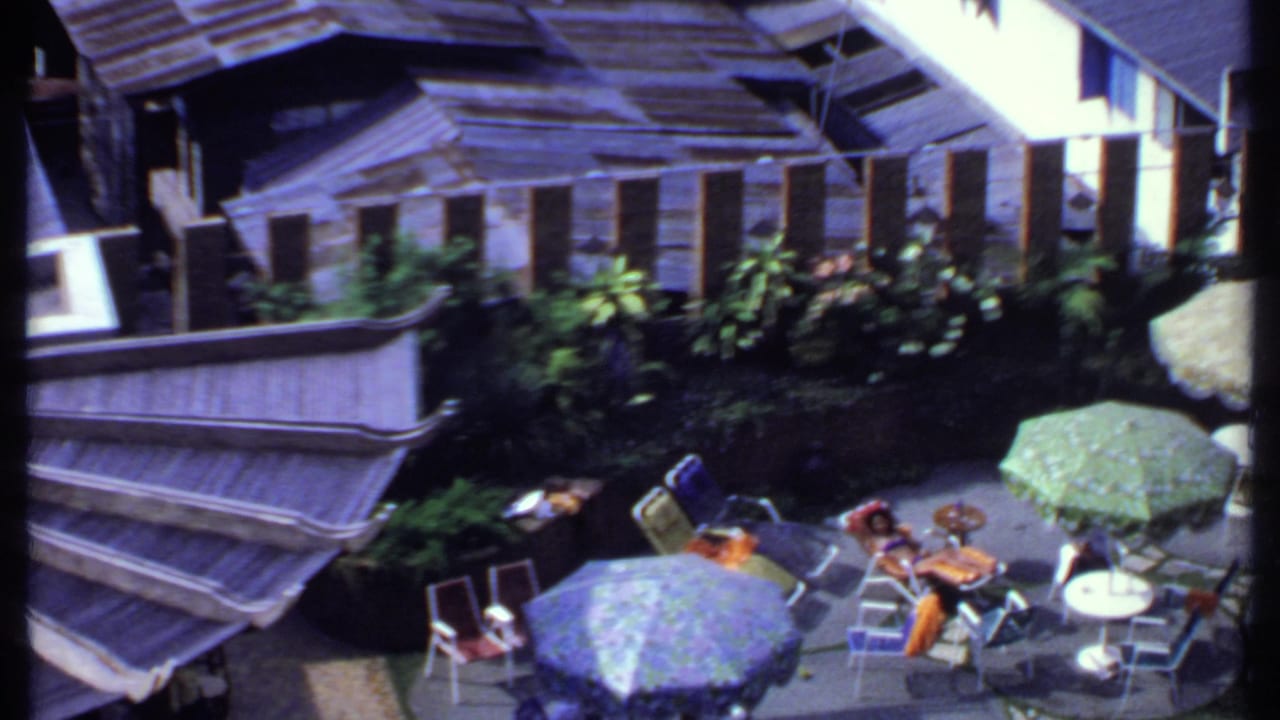 The 1969 India home movie pans across a terrace with multiple parasols, deck chairs, tables, foliage, and traditional roof forms.