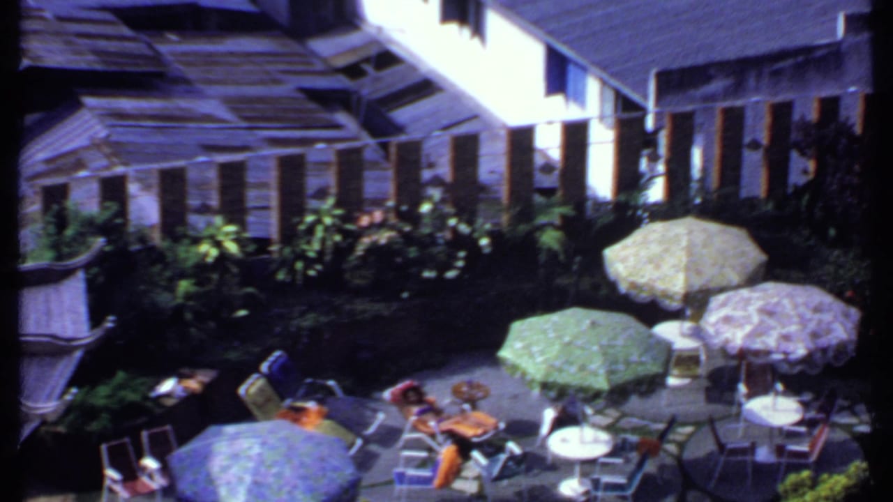 High-angle view of a sunny patio with patterned umbrellas, tables, chairs, greenery, and people seated in shade.