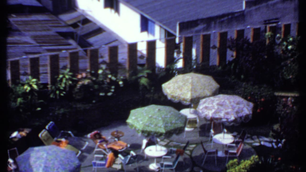 1969 India footage frame with yellow, green, and purple umbrellas over patio seating, garden foliage, and partially visible guests at a backyard gathering.