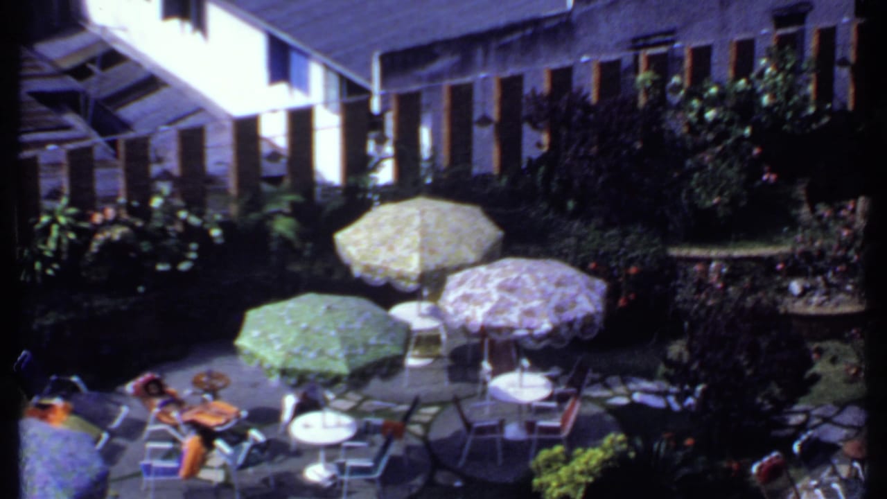 High-angle 1969 India home-movie view of a backyard patio arranged for tea or socializing, with umbrellas, chairs, tables, plants, and seated guests.