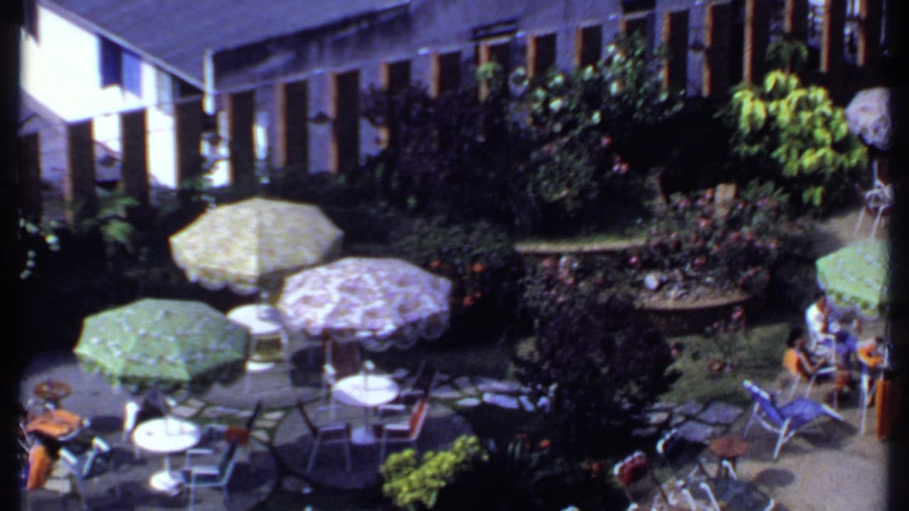 1969 India home-movie frame showing the patio gathering from above, with shaded seating, dense plants, patterned umbrellas, and guests near the garden edge.